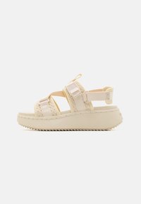 Beige synthetic sandals with woven textile straps, adjustable hooks, and a thick, textured sole featuring a tread pattern for grip.