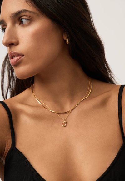 Γυναίκα με μακριά σκούρα μαλλιά που φοράει χρυσούς κρίκους στα αυτιά και στρωματοποιημένα χρυσά κολιέ, ντυμένη με μαύρο αμάνικο τοπ, κοιτάζοντας προς τα αριστερά.