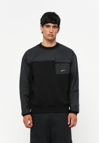 Hanorac negru dintr-un material texturat, având un panou superior de contrast și o buzunar pe piept. Manșete și tiv din tricot.