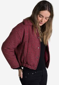 Chaqueta de plumas acolchada a cuadros rojos y negros con cuello tipo camisa, botones de presión, dobladillo fruncido y bolsillos laterales. Llevada sobre una camiseta negra.