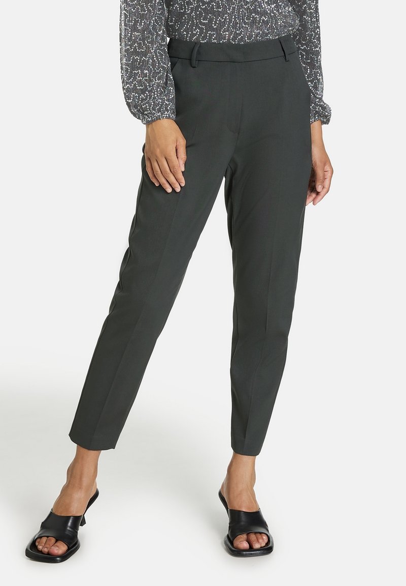 Femme portant un pantalon noir fuselé, un chemisier à manches longues à motifs et des talons ouverts noirs sur fond blanc.