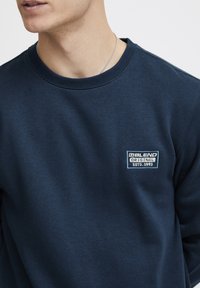 Navyblauwe sweater van zacht materiaal met een ronde hals en een geborduurd logo-patch op de linker borst in wit en lichtblauw.