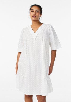 Kvinde, der bærer en hvid knælængde kjole med korte pufærmer, V-udskæring og delikate blondedetaljer.