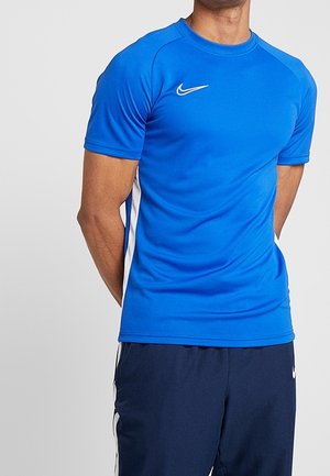 Mężczyzna ubrany w niebieską sportową koszulkę Nike z krótkim rękawem z białymi bocznymi panelami oraz ciemnoniebieskie spodnie dresowe z białymi paskami.