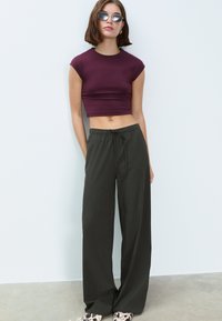 Donkergroene broek met wijde pijpen en een elastische tailleband, gecombineerd met een aansluitende bordeauxrode crop top. Model draagt ronde zonnebril.