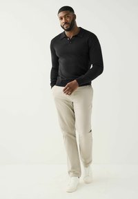 La polo nera a maniche lunghe presenta un colletto e bottoni, abbinata a pantaloni beige slim-fit e sneakers bianche su uno sfondo chiaro.