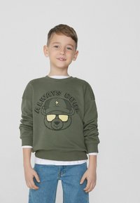 Zelená mikina s grafikou medvěda, který nosí sluneční brýle, text "VŽDY COOL" nahoře. Měkký materiál, kulatý výstřih, volný střih, nošena přes bílé tričko.