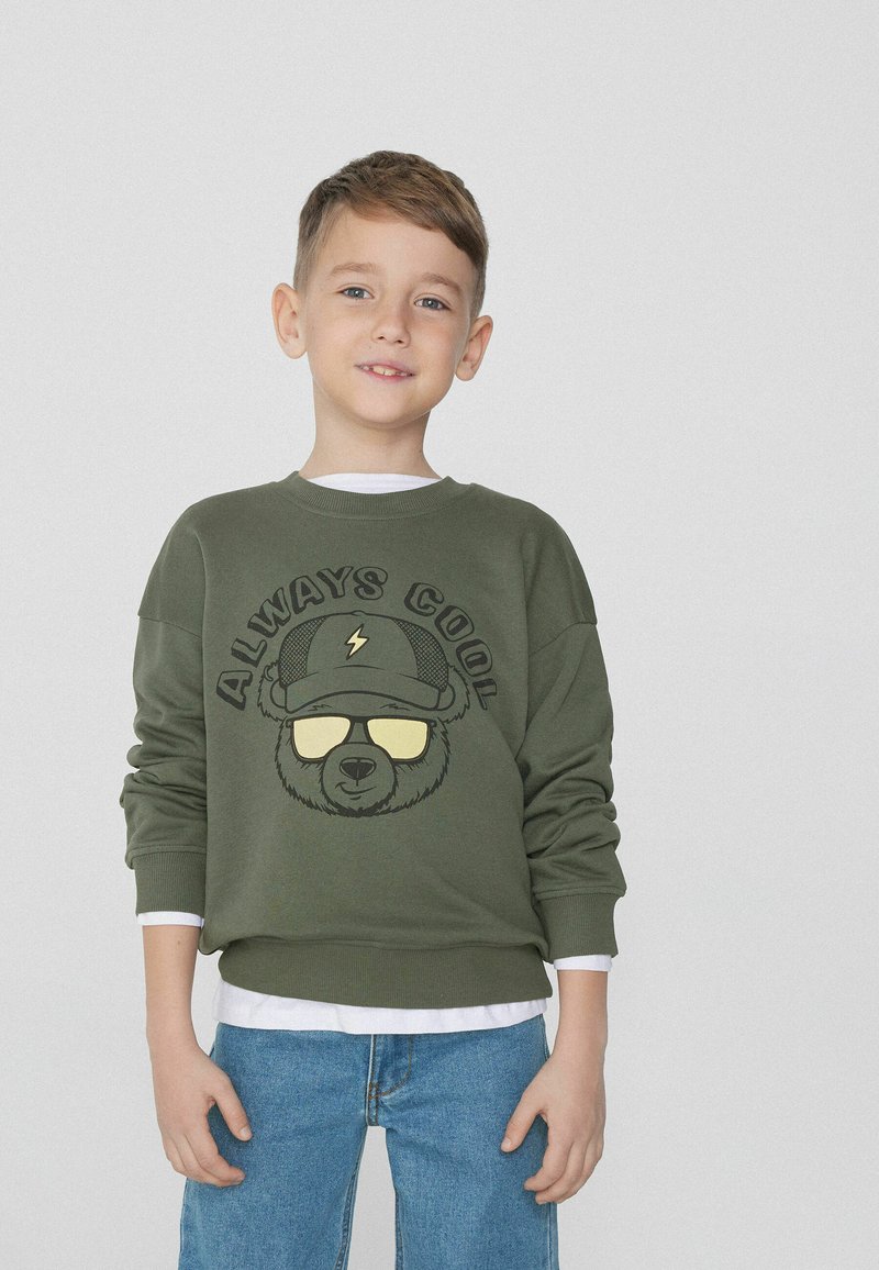 Zelená mikina s grafikou medvěda, který nosí sluneční brýle, text "VŽDY COOL" nahoře. Měkký materiál, kulatý výstřih, volný střih, nošena přes bílé tričko.