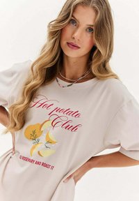 Krátký rukáv béžové bavlněné tričko s nápisem "Hot Potato Club" a grafikou žlutých brambor s bylinkami. Volný střih a měkká textura.