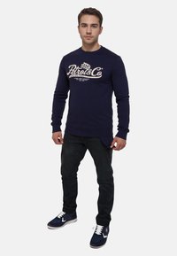 Camicia a maniche lunghe blu marina con logo e testo bianchi, abbinata a jeans scuri e scarpe sportive blu marina con lacci bianchi e dettagli viola.