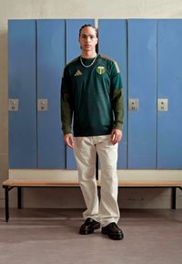 Groen sportshirt met horizontale strepen, gouden accenten en teamlogo. Gecombineerd met witte broek en zwarte chunky schoenen, voor blauwe lockers.