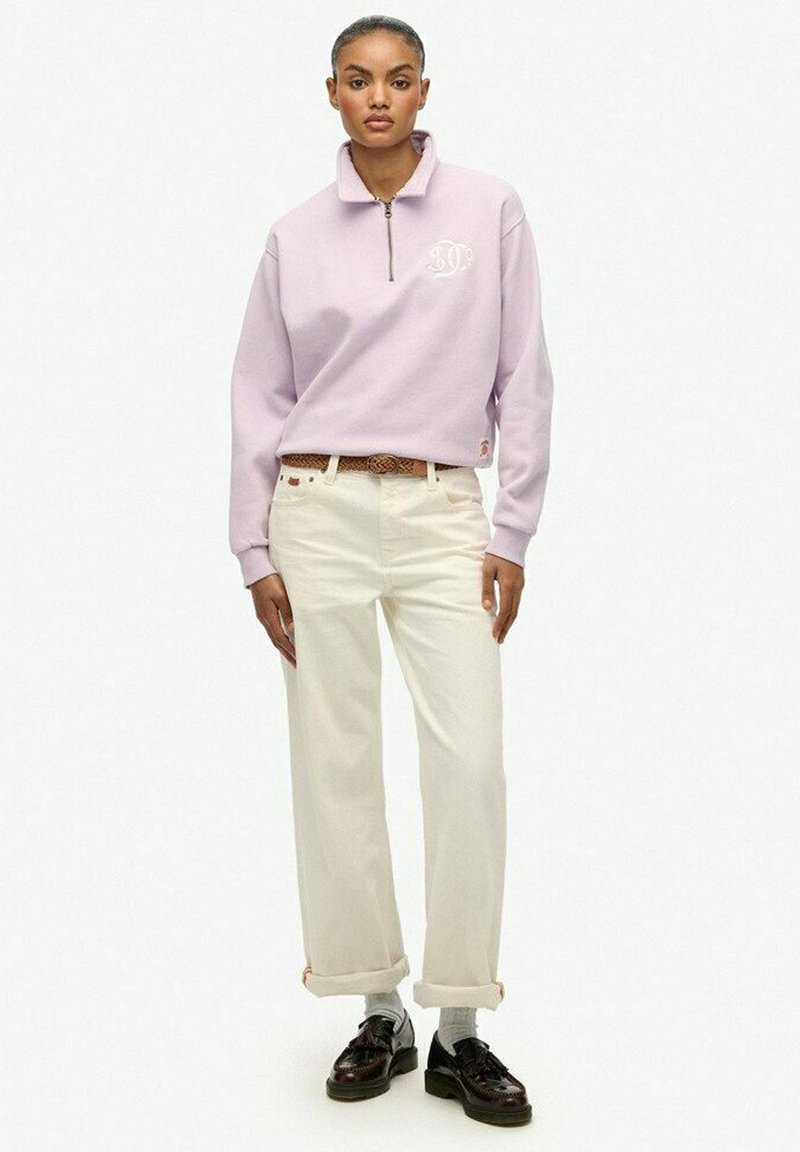 Kobieta ubrana w jasnofioletowy sweatshirt z ćwierć zamkiem, kremowe spodnie o szerokich nogawkach z brązowym paskiem, białe skarpetki i ciemnobrązowe mokasyny stoi na tle białym.