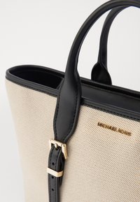 Beige kudotettu kangaskassi mustilla nahkaaksenteilla ja -kahvoilla. Varustettu kultasävyisellä logolla ja soljella hihnassa.