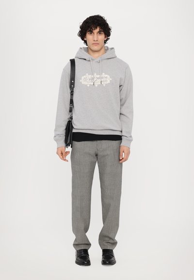 Grauer Kapuzenpullover mit cremefarbenem Druck, über einem schwarzen Shirt getragen. Kombiniert mit karierten grauen Hosen und schwarzen Schuhen. Schwarze Tasche über der Schulter.
