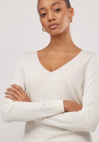 Cremefarbener V-Ausschnitt-Pullover aus leichtem, glattem Material mit langen Ärmeln. Gerippte Bündchen und ein klares Design, das eine minimale Ästhetik zur Schau stellt.