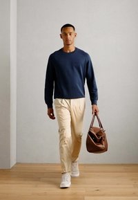 Navy sweatshirt, cream pants, and white sneakers worn by a man carrying a brown leather bag. Smooth textures and relaxed fit are evident.