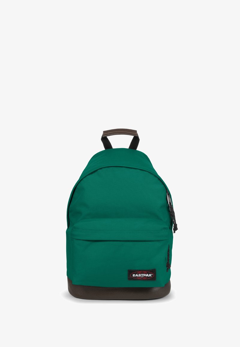 Mochila de tela verde con forma redondeada, que incluye un bolsillo frontal y detalles marrones en la parte inferior. Cuenta con correas negras ajustables y un parche con el logo.