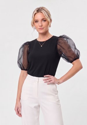 Femme aux cheveux blonds portant un haut noir à manches bouffantes transparentes et un pantalon blanc taille haute, posant devant un fond clair.