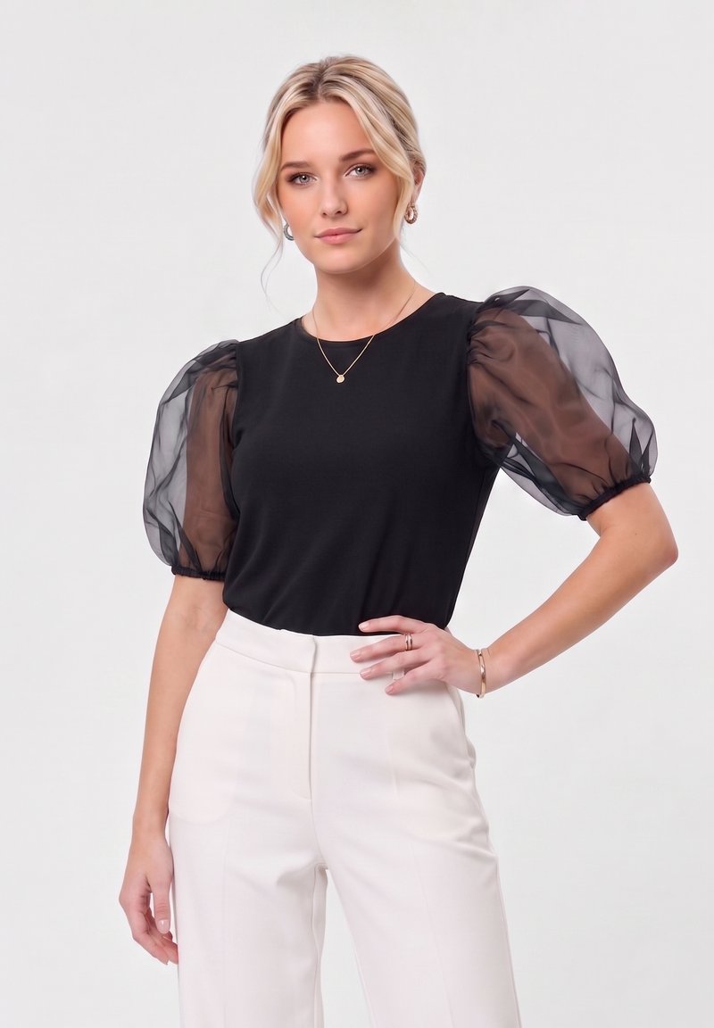 Femme aux cheveux blonds portant un haut noir à manches bouffantes transparentes et un pantalon blanc taille haute, posant devant un fond clair.