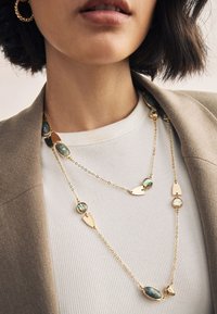 Gold-tone layered necklace featuring various oval and teardrop-shaped accents with iridescent stones against a textured white top.