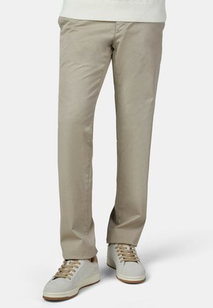 Man wearing beige straight-leg pants and white sneakers with beige laces, standing on a white background.