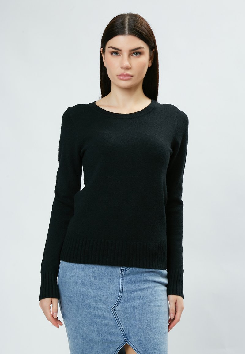 Pull en tricot noir avec un col rond et des manches longues, présentant des détails côtelés au niveau de l'ourlet. Porté par-dessus une jupe en denim bleu clair.