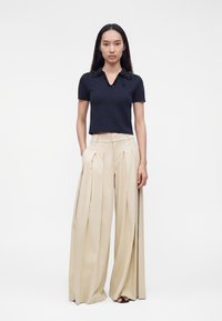 Femme aux longs cheveux noirs portant un haut noir à manches courtes avec col et un pantalon beige plissé, large et taille haute, les mains dans les poches.