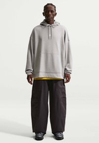 Jeune homme aux cheveux tressés portant un sweat à capuche gris surdimensionné Nike, une chemise jaune, un large pantalon cargo noir et des baskets noires, debout devant un fond blanc.