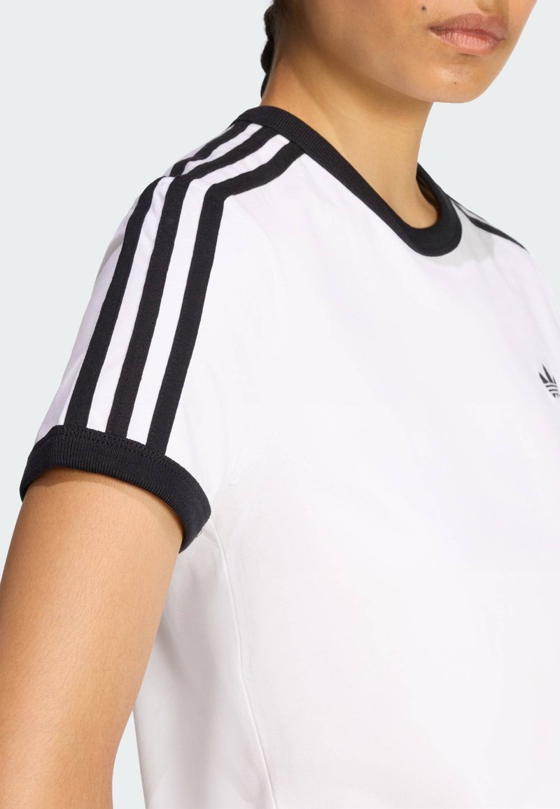 Hvid T-shirt med sort rund halsudskæring og kontrasterende sorte ærmekanter. Har tre sorte striber ned langs hver skulder.