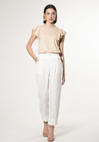 Top beige con maniche a volant, abbinato a pantaloni bianchi con dettagli di bottoni dorati e risvolti. Modella che indossa tacchi nudi.