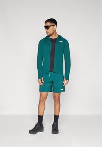 Haut à fermeture éclair teal sur une chemise noire, assorti à un short teal. Baskets noires avec des semelles texturées et des chaussettes sombres. Des lunettes de soleil complètent le look.