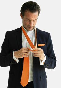 Corbata naranja siendo anudada, combinada con un traje azul marino y una camisa blanca. Detalles notables incluyen un pañuelo de bolsillo a juego y gemelos.