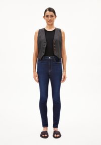 Gilet en denim gris avec des accents en bouton, haut noir ajusté, jeans skinny bleus et sandales à enfiler noires. Le mannequin se tient contre un fond blanc.