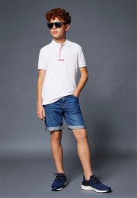 Hvit polo-skjorte med røde detaljer, dongerishorts rullet ved kanten, og marineblå joggesko med teksturert overdel. Svart solbriller fullfører looken.