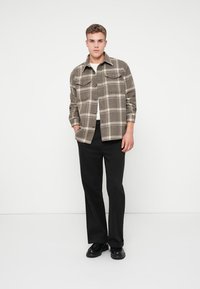 Grijze geruite shirt met een kraag, knoopsluiting aan de voorkant en twee borstzakken, gecombineerd met zwarte broek en zwarte schoenen.