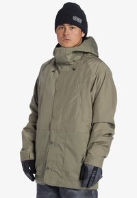 Khaki sneajakke med høj krave, flere trykknapper og to frontlommer. Sort strikket hue og handsker supplerer outfittet.