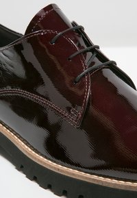 Chaussure à lacets en cuir verni bordeaux avec lacets noirs, couture texturée, bande beige et semelle en caoutchouc noire à crampons profonds.