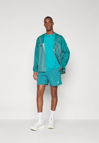 Teal lichtgewicht jasje met grijze accenten, gecombineerd met een gestructureerd teal shirt en shorts. Witte sportschoenen en crew sokken maken de look compleet.