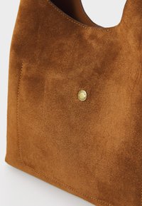 Brown suede handbag with a smooth surface and a small gold circular logo. It features a curved top opening and a visible pocket on the side.