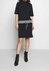 Vestido negro con mangas cortas y escote redondo, que presenta una cinturilla a rayas y un detalle atado. La modelo lleva botines negros.
