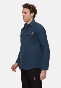 Camicia da uomo in flanella blu con abbottonatura davanti, due tasche sul petto e bottoni marroni. Presenta una texture morbida e un colletto.