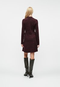 Abito bordeaux con maniche lunghe, tessuto a maglia testurizzato, che arriva sopra il ginocchio. Abbinato a stivali neri alti fino al ginocchio.