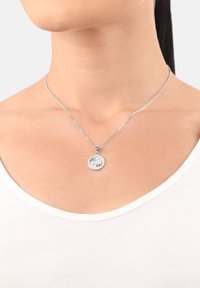 Silverhalsband med en cirkulär berlock med ingraverade detaljer, p�å en tunn kedja som vilar mot en vit skjortkrage.