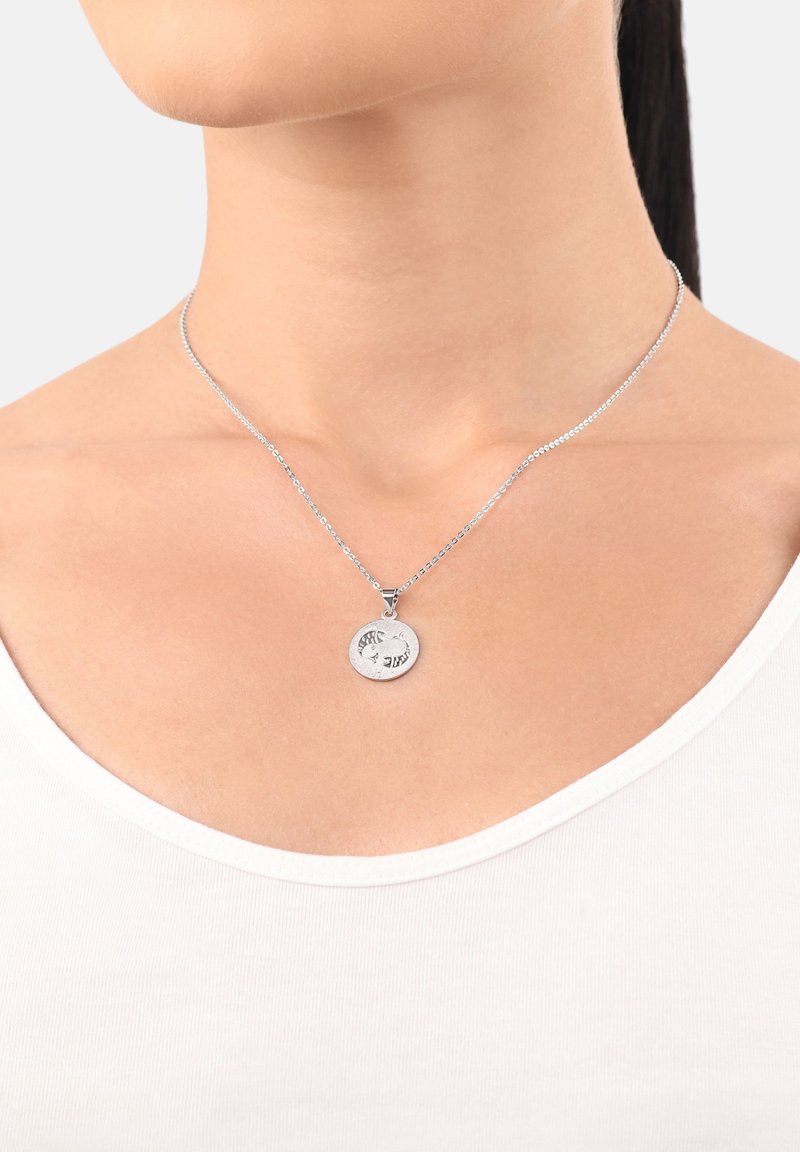Silverhalsband med en cirkulär berlock med ingraverade detaljer, på en tunn kedja som vilar mot en vit skjortkrage.