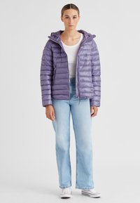 Lila Puffjacke mit Kapuze, ausgestattet mit einem horizontal gesteppten Design und Reißverschlusstaschen, kombiniert mit hellblauen Schlagjeans und weißen Sneakers.