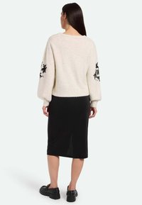 Helle Cremefarbene Pullover mit schwarzem floralem Stickmuster auf Ballonärmeln, getragen über einem figurbetonten schwarzen Midirock, kombiniert mit schwarzen Plateauschuhen.