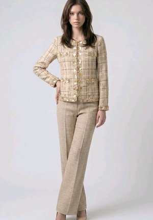Mujer con chaqueta texturizada de cuadros beige con botones dorados y pantalones beige de pierna ancha a juego, de pie con una mano en la cadera.
