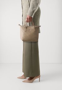 Bolso acolchado beige con dos asas superiores y logotipo metálico, combinado con una falda larga verde oliva y tacones en color nude.