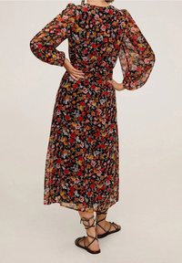 Femme portant une robe midi noire à fleurs avec des manches longues transparentes et bouffantes, se tenant les mains sur les hanches, le dos tourné vers l'appareil photo.
