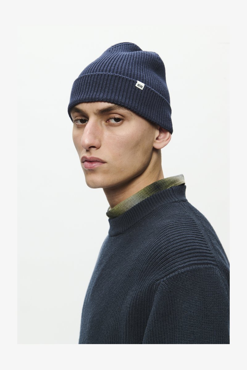 Gorro de malha canelada azul-escuro com um pequeno rótulo branco. Usado com um suéter escuro e uma gola de camisa xadrez verde visível por baixo.